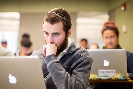 一个戴圆顶小帽的男学生的照片, 坐在波胆网站的演讲厅里，看着他的电脑