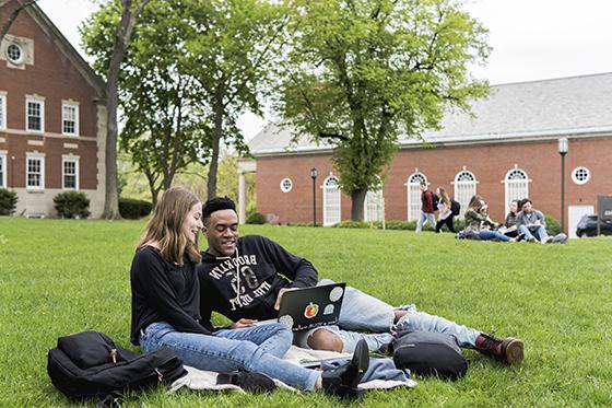 Two Chatham University students sit on a green grass academic quad, working toge的r on a laptop. 