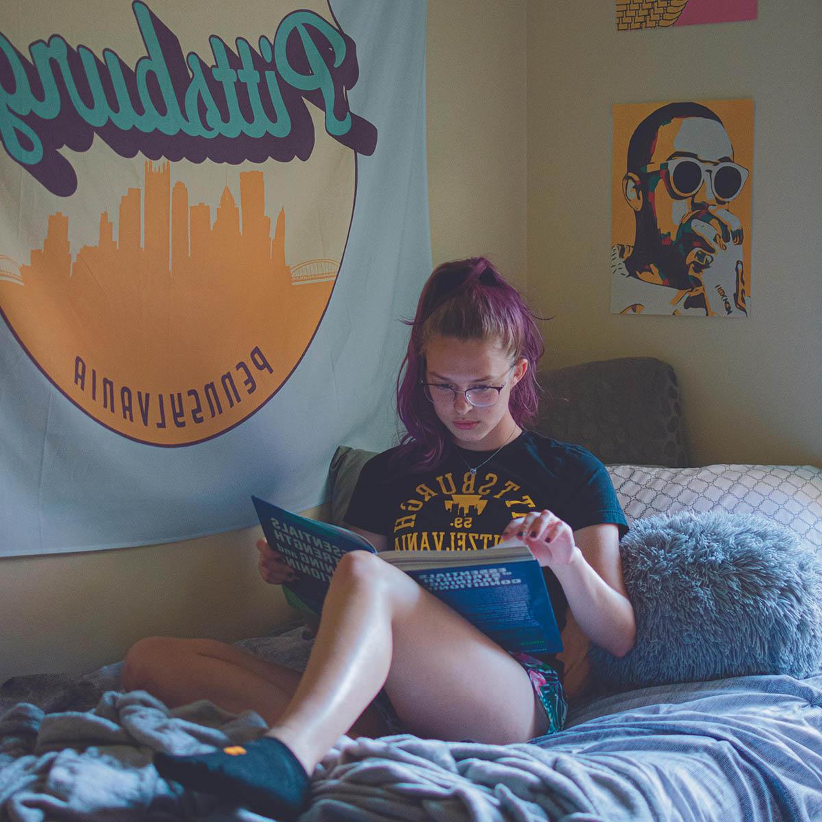 Photo of a young female student with a pink ponytail and glasses reading on her dorm room bed. A large tapestry that says Pittsburgh, Pennsylvania is on the wall