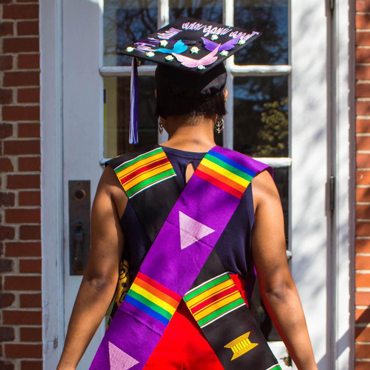一个学生背对镜头的照片, 穿着彩虹色的毕业披肩，戴着一顶写着“这些翅膀是为飞翔而生”的毕业帽。